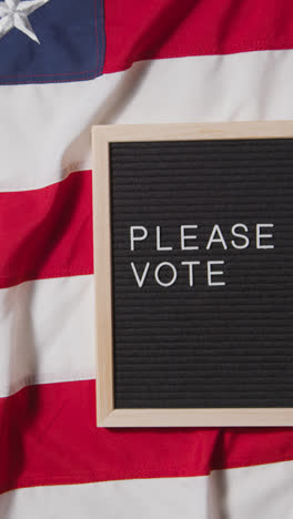 Vertical-Video-Of-Sign-Reading-Please-Vote-Lying-On-US-Stars-And-Stripes-Flag-For-American-Election-2024-2